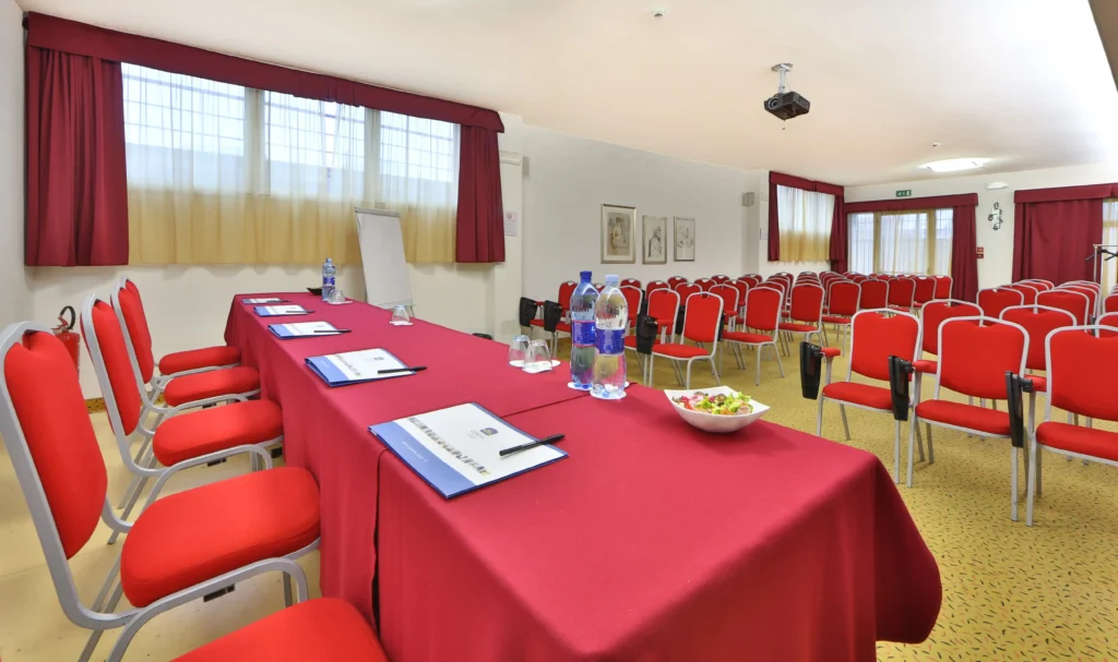 In the picture, the Romea meeting room of the Cesena Hotel: we can see the speakers' table with notebooks, pens and bottles of water. In front of the table there are some rows of chairs. The room is surroundend by windows with closed curtains.