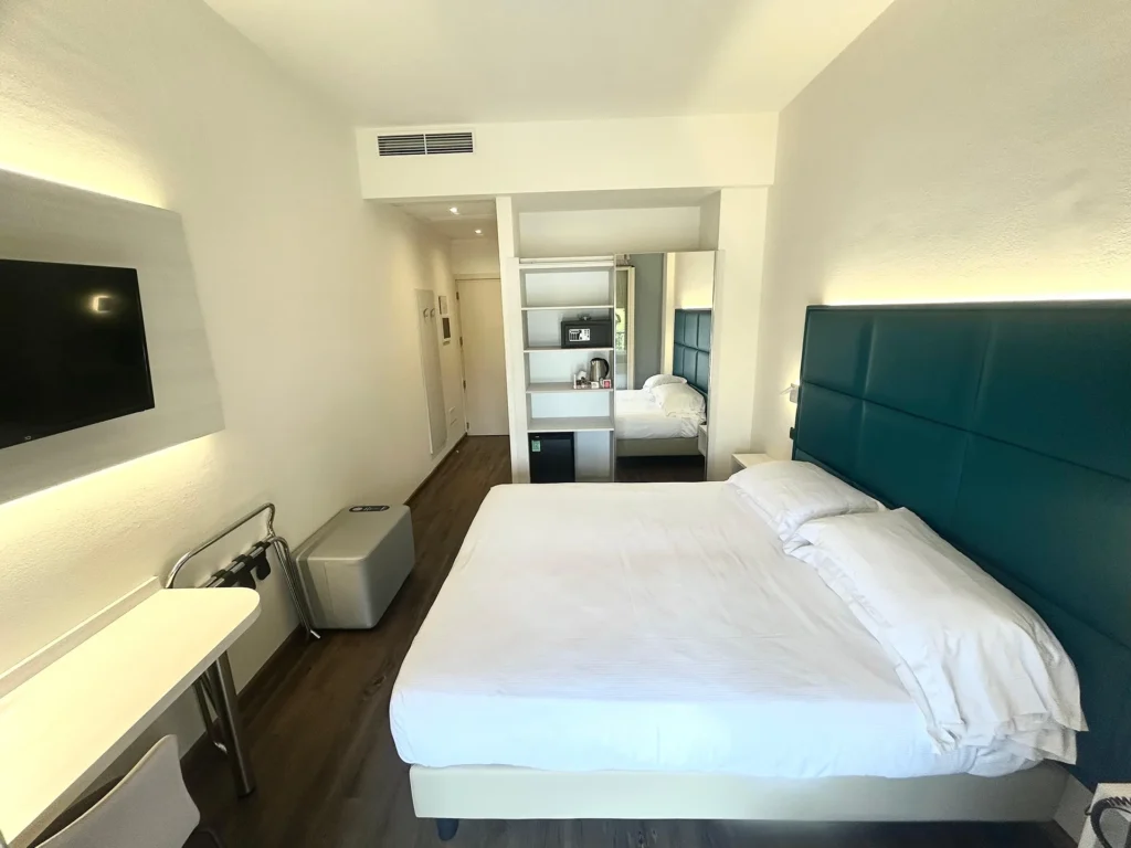 In the picture, the Standard Room of the Cesena Hotel: we can see the double bed and, opposite, a small table with a chair and a TV hanging on the wall. On the right of the bed, there is a full-length mirror and shelves with a kettle, safe, and minibar.